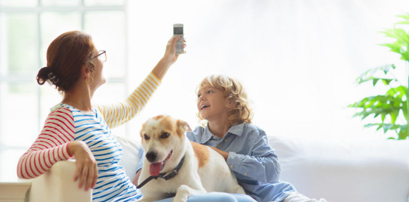 A woman operating the remote control for an air conditioning unit while seated on a couch with a child and a dog.